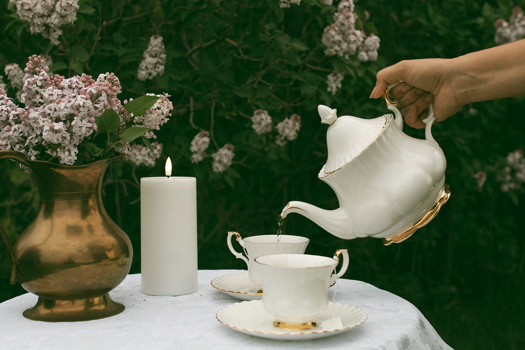Sundays Tea being poured from a white teapot into a cup on a table with flowers and a candle.