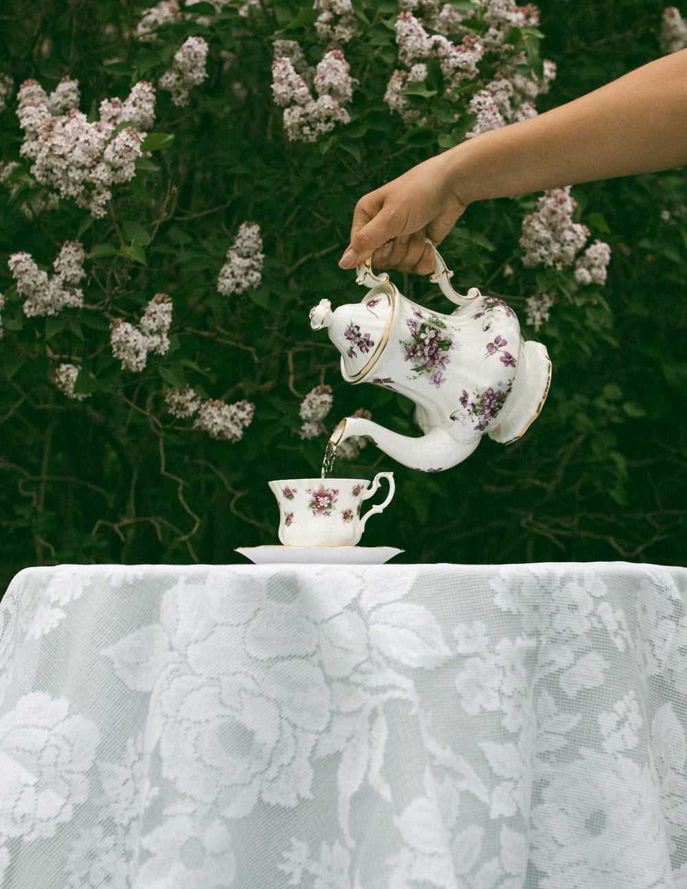 Person pouring Sundays Tea, and organic and Canadian tea, from a decorative teapot into a matching cup on a floral tablecloth.